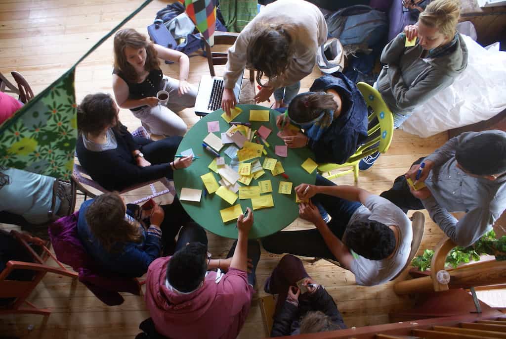 A birds eye view shot of 10 individuals sat and stood around a circle table covered in post-it notes and pens.