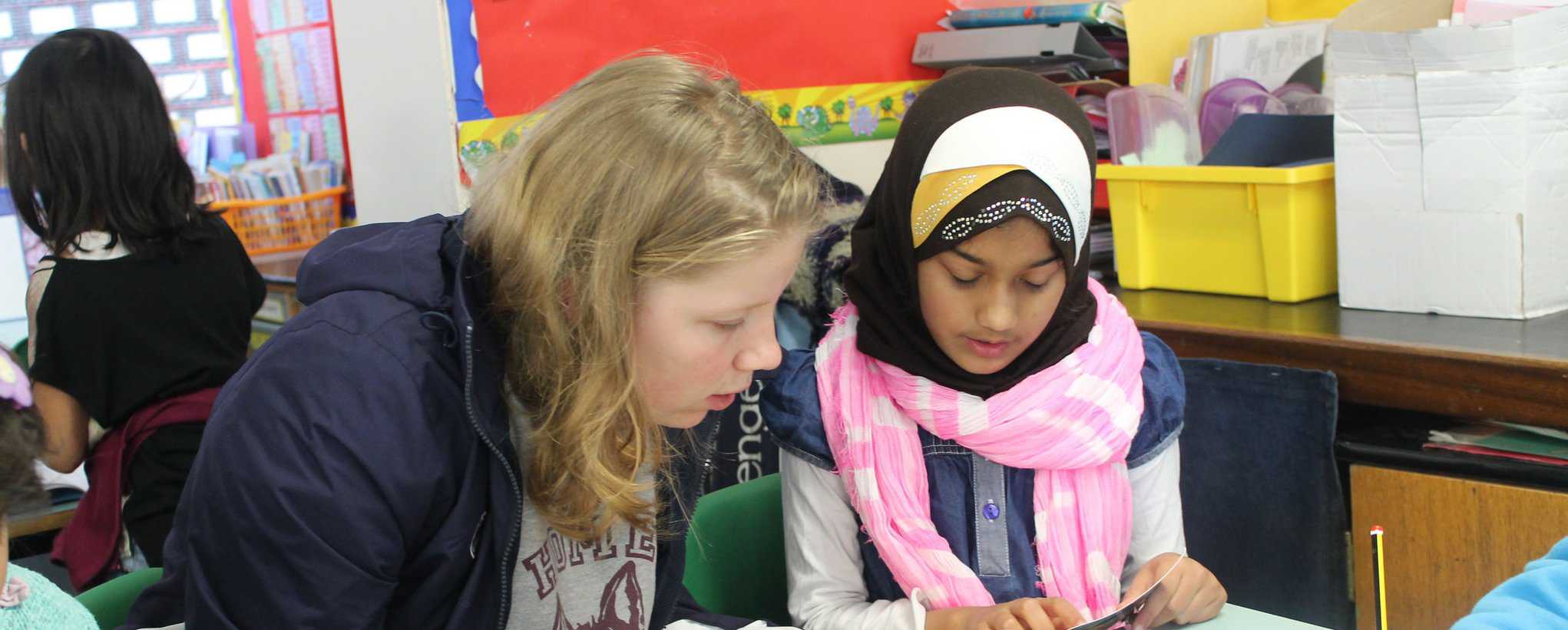 A Schools Plus volunteer sits with a pupil reading. They are in a classroom.