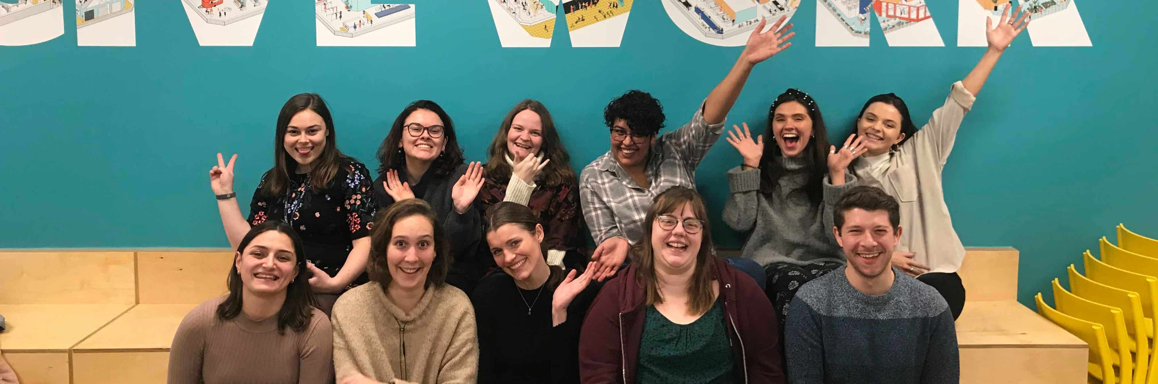 Ten members of the Student Hubs staff team smile at the camera with their arms in the air. They are sat in two rows on a brown step in front of a turquoise wall.