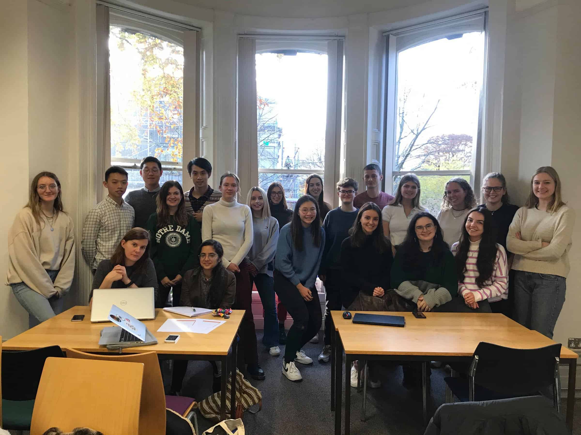 21 people stand in front of a window in a classroom smiling at the camera.