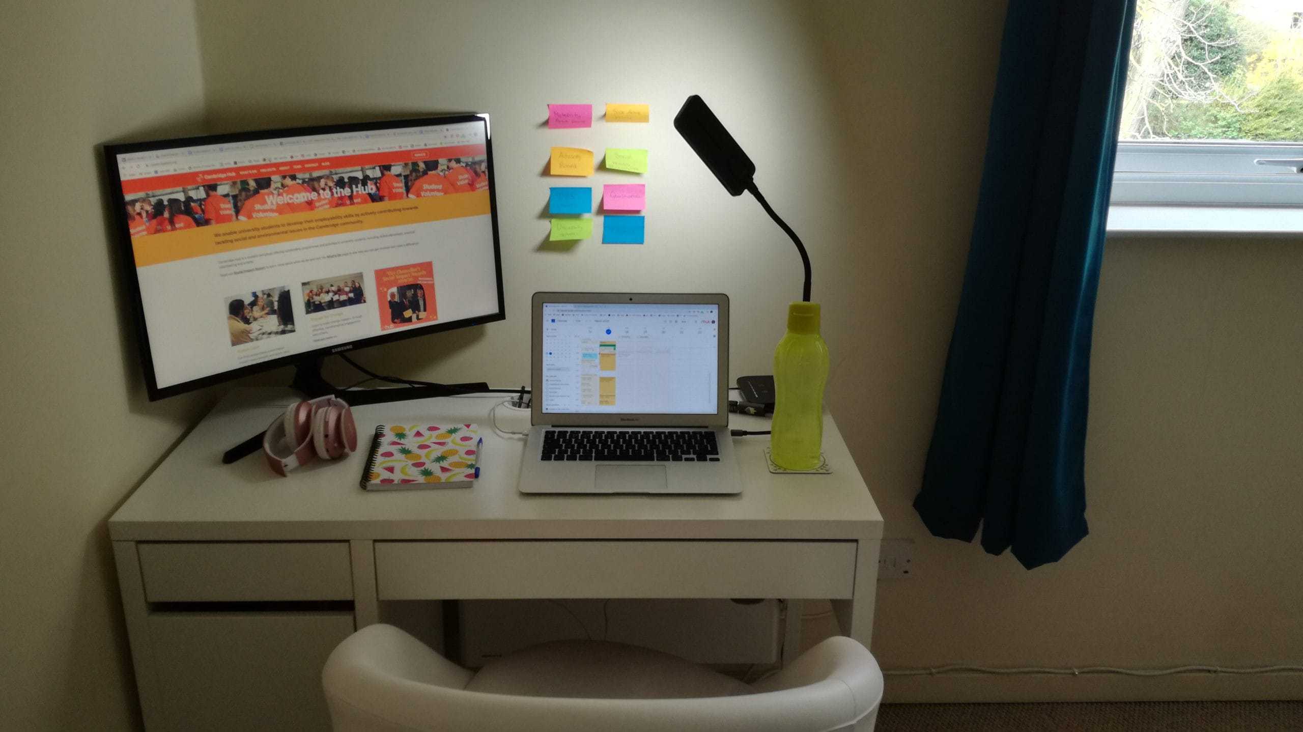 A white desk with a white chair tucked in has a monitor, laptop, pink headphones, notebook and yellow water bottle on it. On the wall are 8 multicoloured post-it notes.