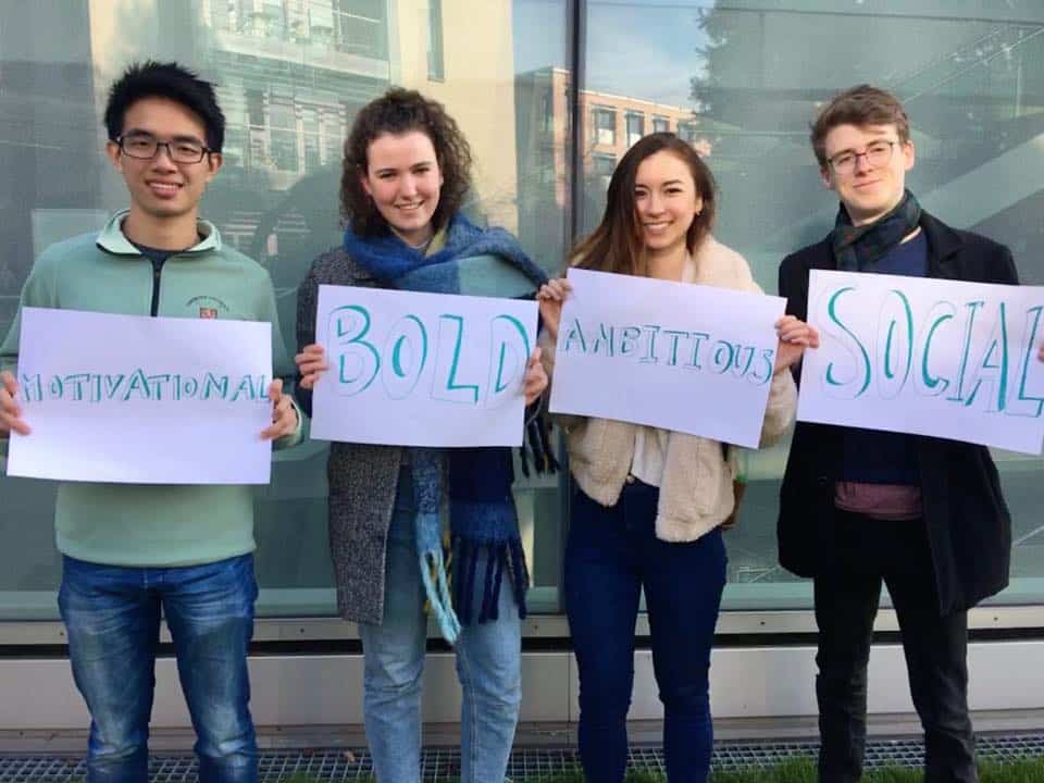 A picture of four students from Cambridge Hub holding up some of the Student Hubs values written on A3 paper. They are in front of a glass shop window. Values are motivational, bold, ambitious and social.