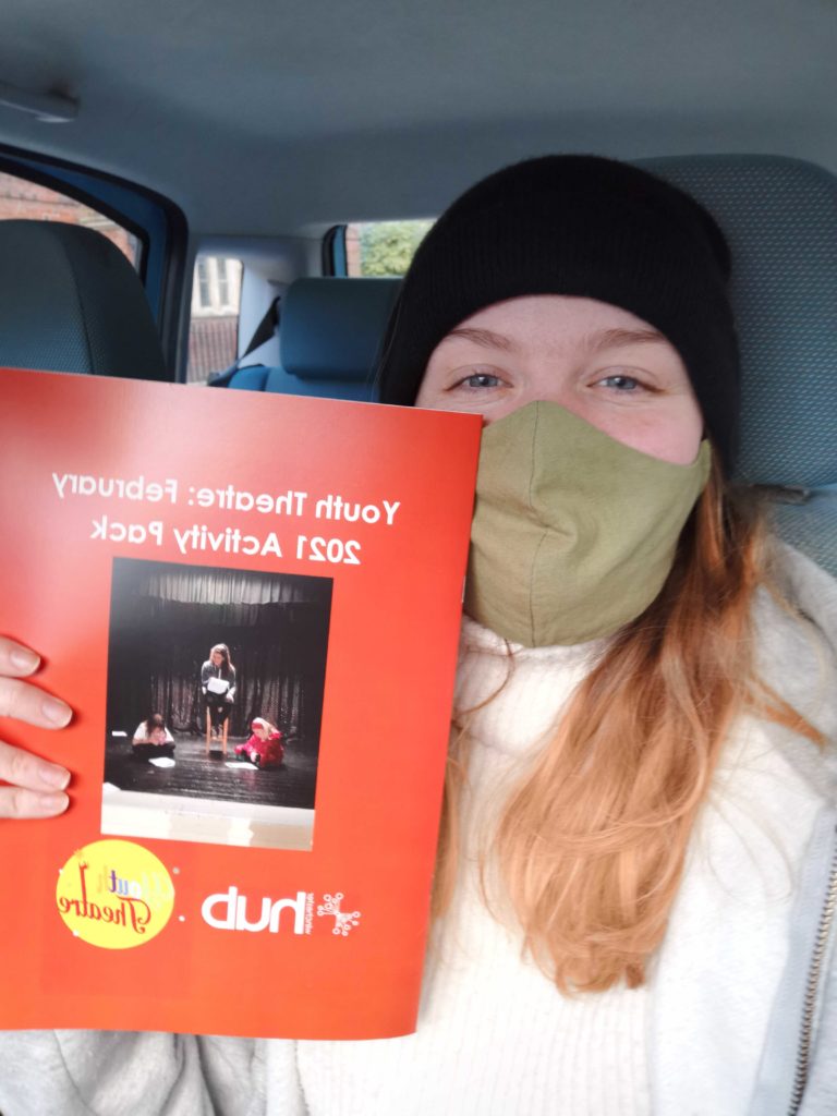 Liz Alcock, Winchester Hub Manager, looks at the camera wearing a green face mask holding up an orange Youth Theatre booklet
