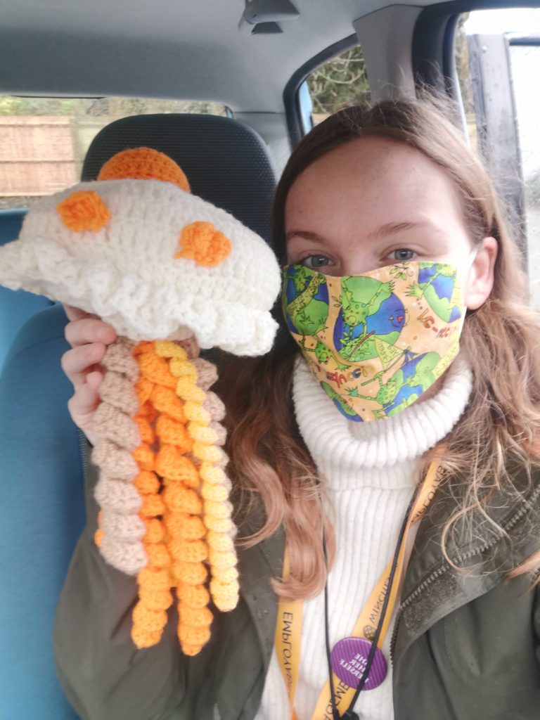 Liz Alcock, Winchester Hub Manager, looks at the camera wearing a colourful face mask holding up an orange Youth Theatre booklet