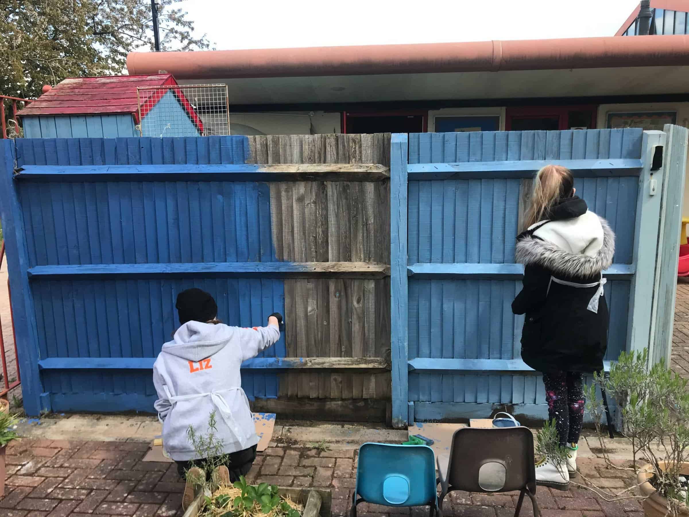 The backs of two volunteers painting a fence in two shades of blue. One is crouching and one is standing.