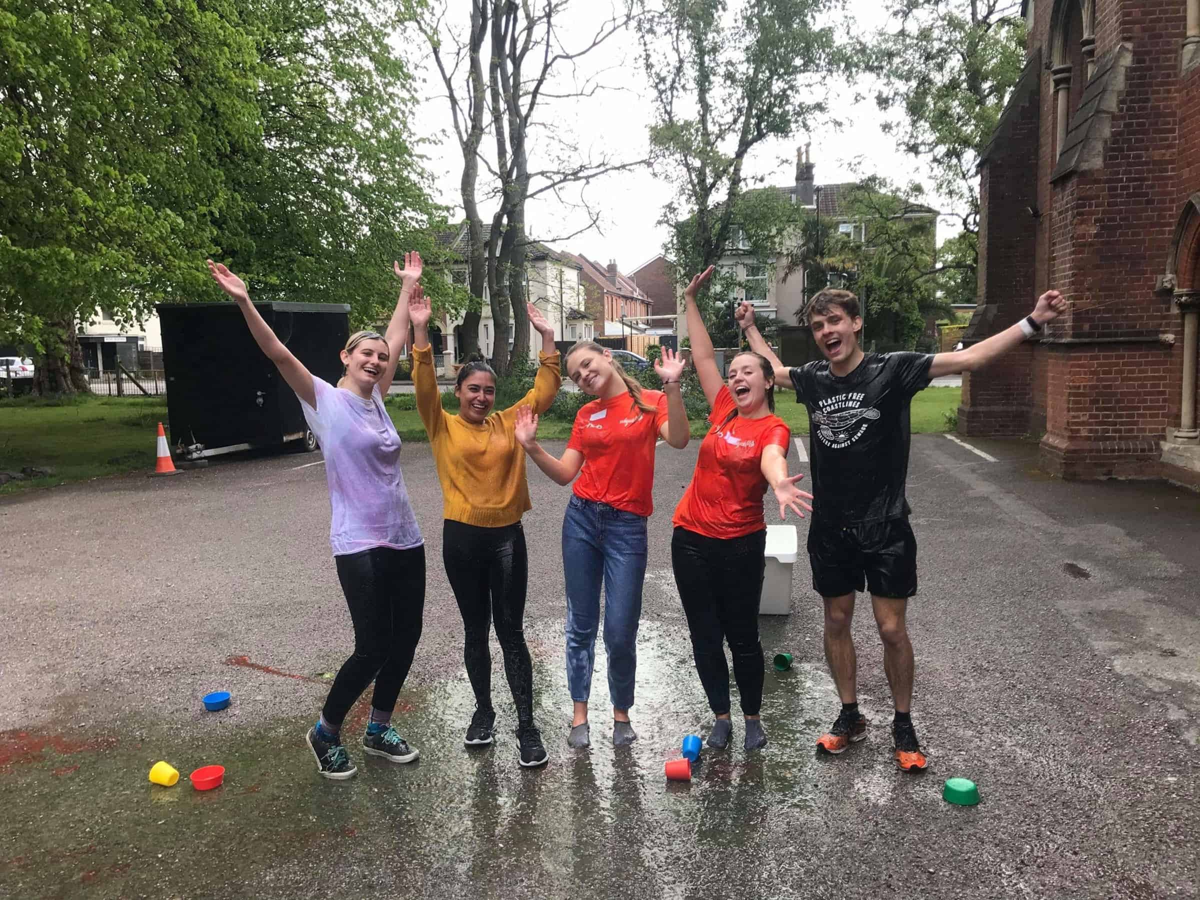 Five students are standing up with their arms in the air smiling. They are wearing wet clothes and stood on tarmac. 