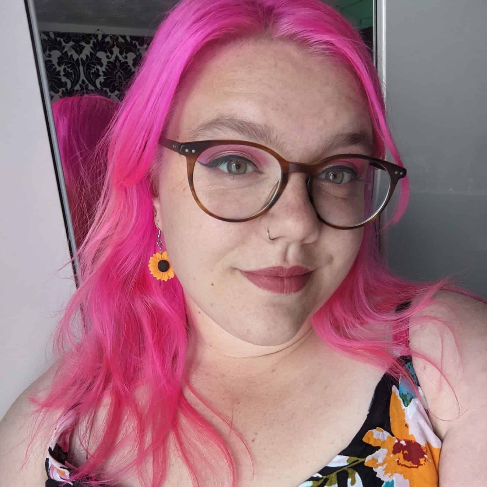 Southampton Hub Programme Manager, Catherine Taplin-Thorpe is smiling at the camera. Catherine has bright pink hair and pink eyeshadow and lipstick. Catherine is wearing sunflower earrings and a floral top. 