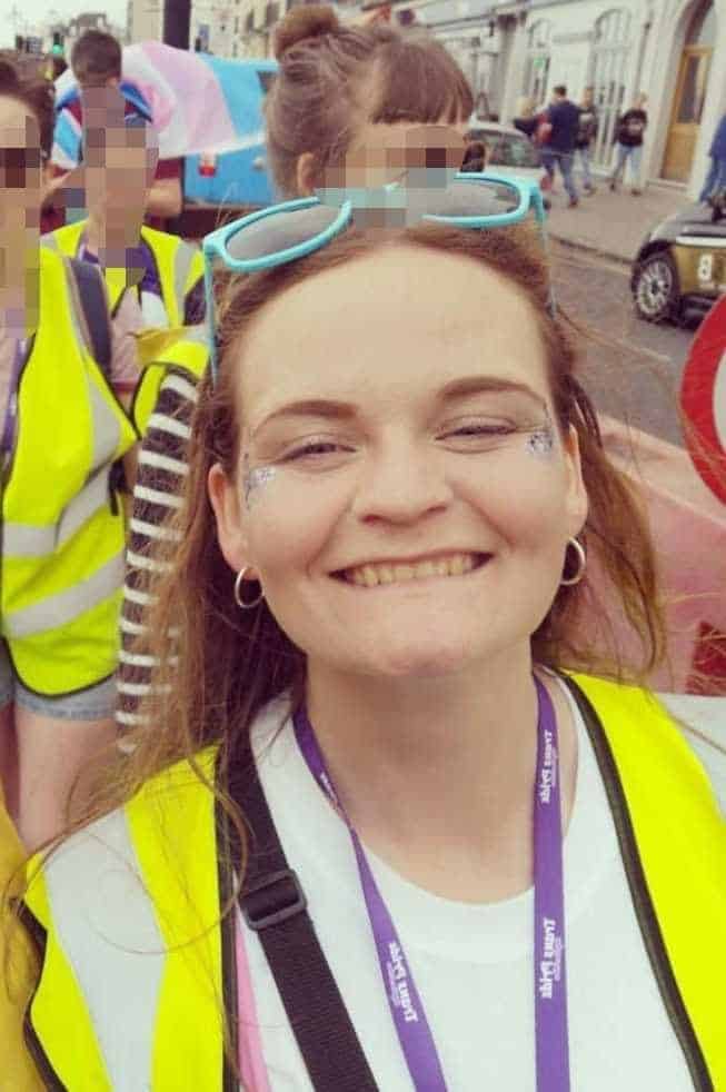 Winchester Hub Manager, Liz Alcock, is smiling at the camera. Liz is wearing a white tshirt, a neon yellow hi vis vest and a purple landyard. Liz has blue sunglasses on their head and glitter by their eyes. In the background is other people in hi vis jackets. 