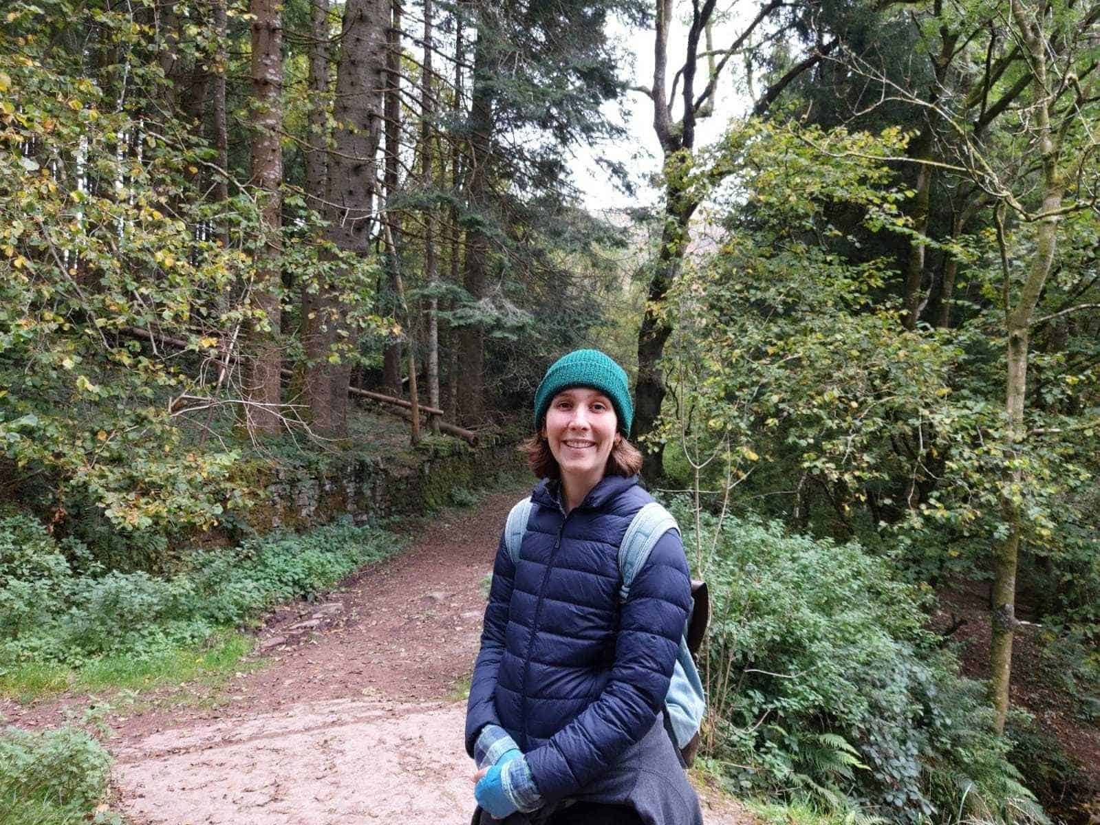 Winchester Hub Programme Manager, Sorcha Young, is standing in woodland. Behind Sorcha is a brown path and a variety of trees and foliage. Sorcha is smiling at the camera wearing a turquoise hat, blue puffer jacket, blue gloves and a blue backpack. 