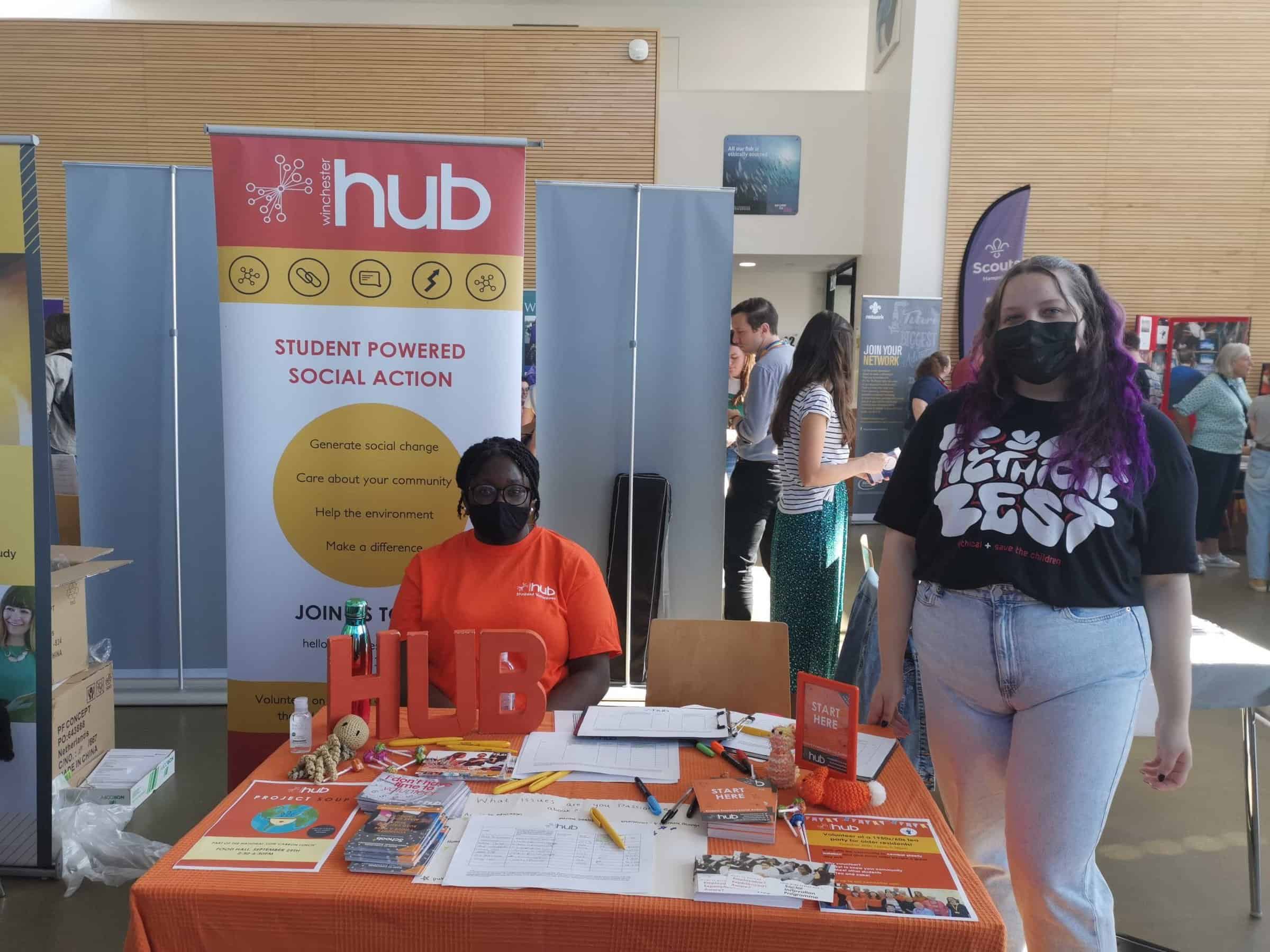 Two volunteers are at a table with an orange table cloth covered in leaflets. One is sat behind the table wearing an orange t shirt, the other is stood to the right of the table wearing a black t-shirt. Behind the table is a banner reading "Winchester Hub".