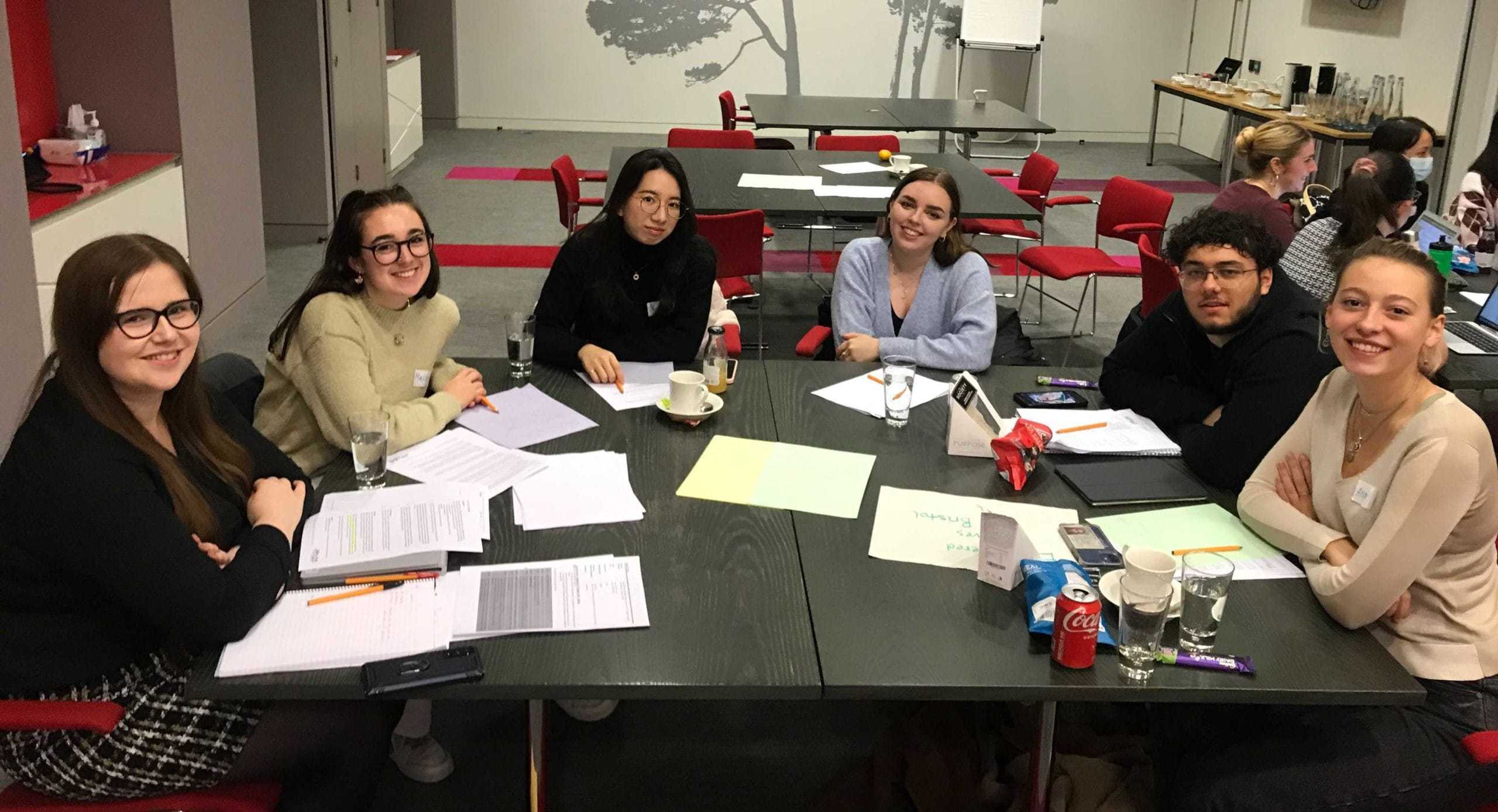 6 students smile at the camera. They are sat at a table with papers on it.