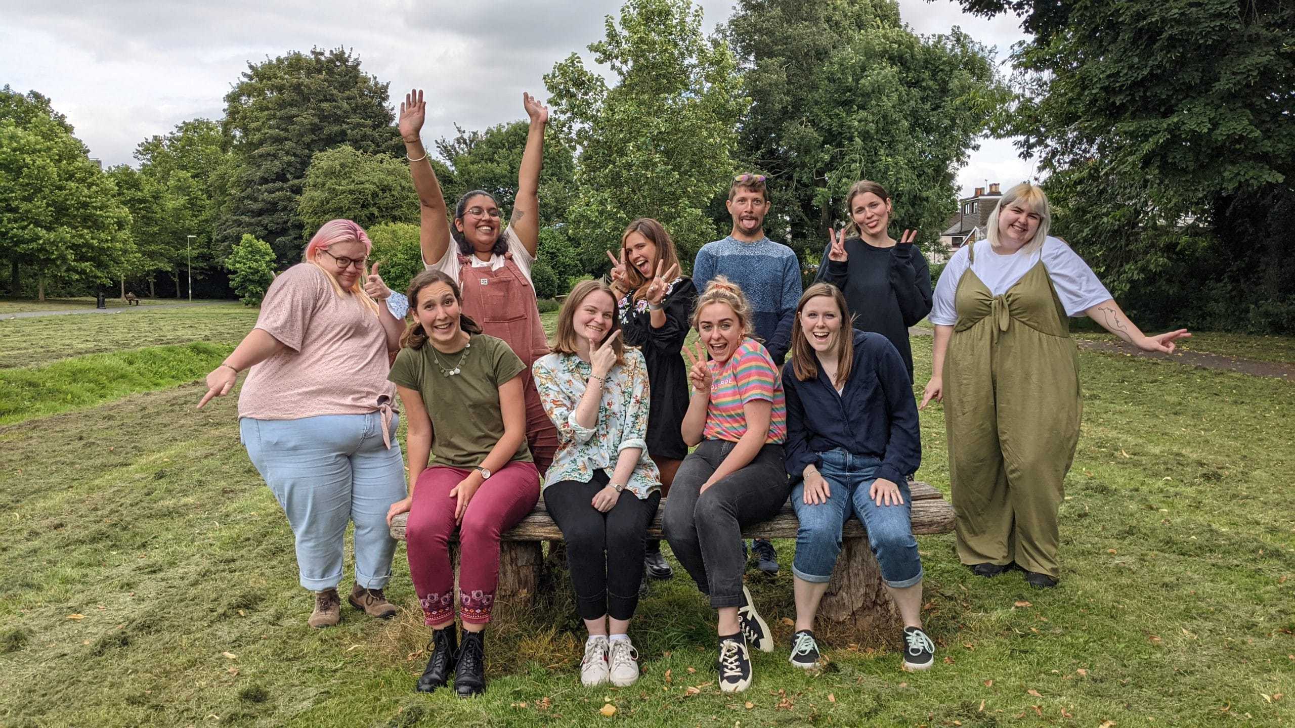 Four members of the Student Hubs staff team sit on a bench, standing behind them is another five members of the team. They are in a park and everyone is looking at the camera pulling a silly face or pose.