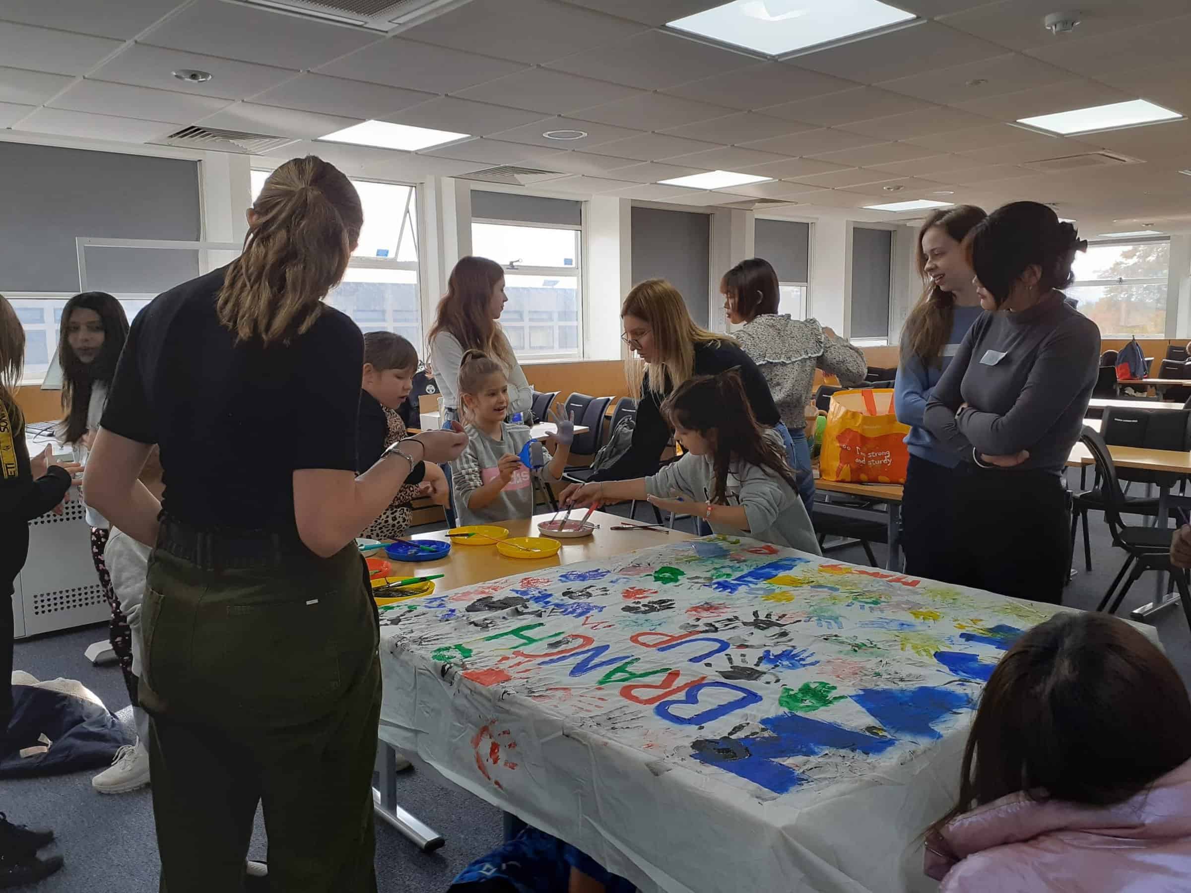 14 students and young people are mingling in a room around a table. On the table a big white cloth reads BRANCH UP in multicoloured paint with painted handprints around it.