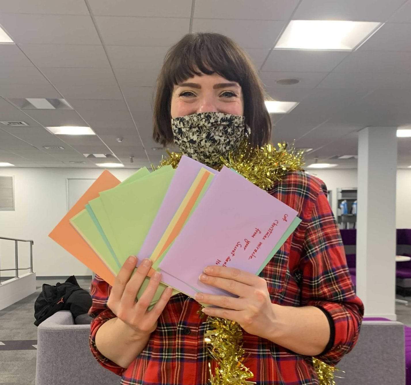 A Kingston Hub volunteer looks at the camera wearing a face mask and a tinsel scarf holding several multicoloured cards