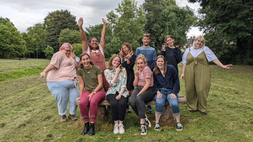 In a park, Student Hubs staff members are making silly faces and throwing their hands in the air. There are four sat down on a bench and six stood behind them.