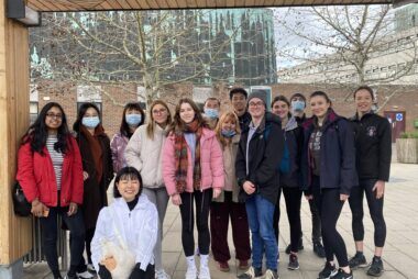 14 students smile at the camera wearing coats and standing outdoors underneath a wooden shelter