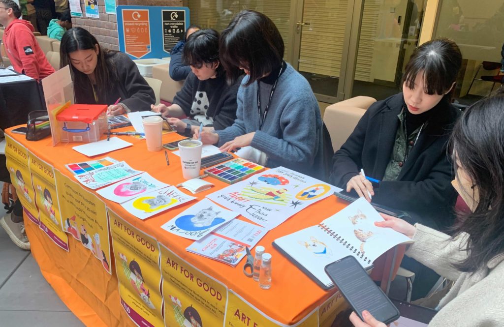 Five students sit around a table drawing. The table has an orange tablecloth and is covered in illustrations. 