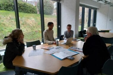 4 people sit at a wooden table in conversation. In the background is a large window overlooking grass.