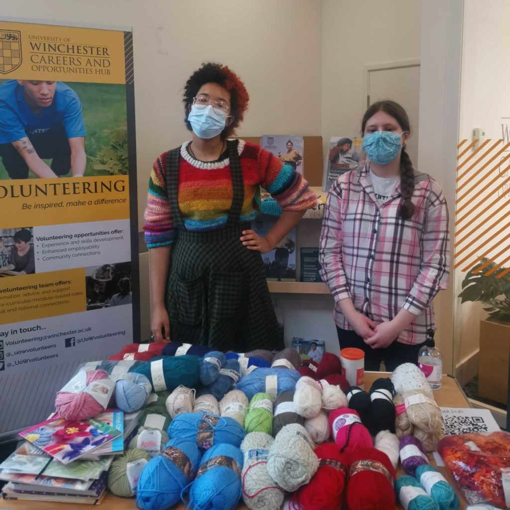 2 students stand behind a table covered in yarn. 