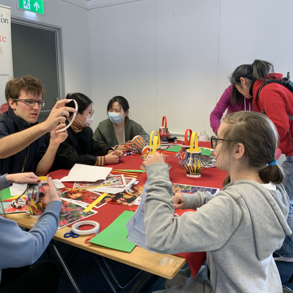 7 young people and students sit around a table making crafts. On the table is a red table cloth and a range of paper, scissors and tape.