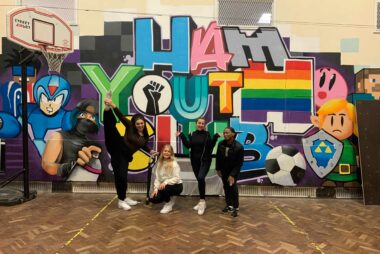 four individuals look at the camera striking a pose. They are in front of colourful graffiti that reads "Ham Youth Club"