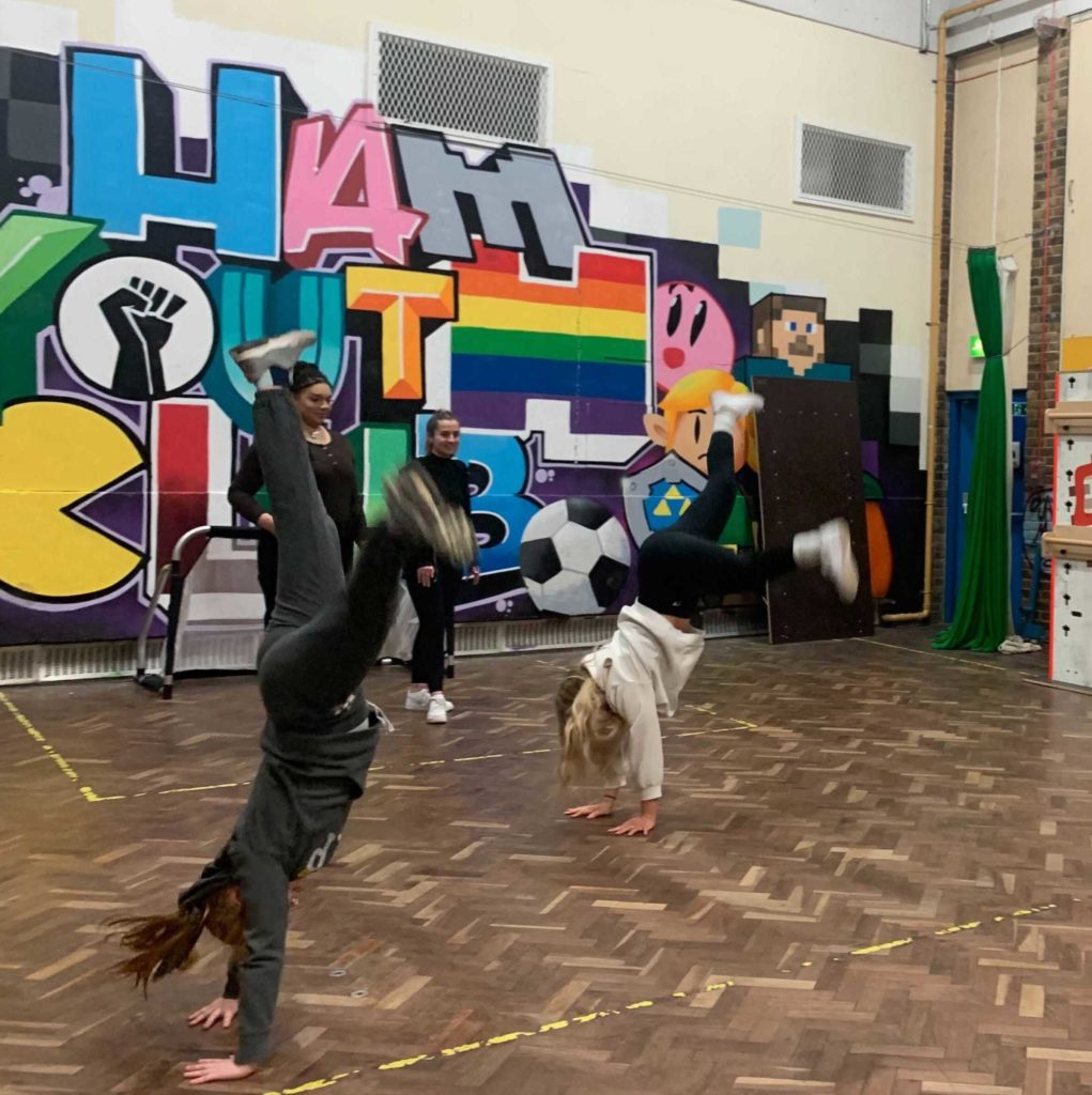 Two young people do cartwheels in a sports hall whilst two students volunteers stand watching. The background of the shot is graffiti reading "Ham Youth Club"