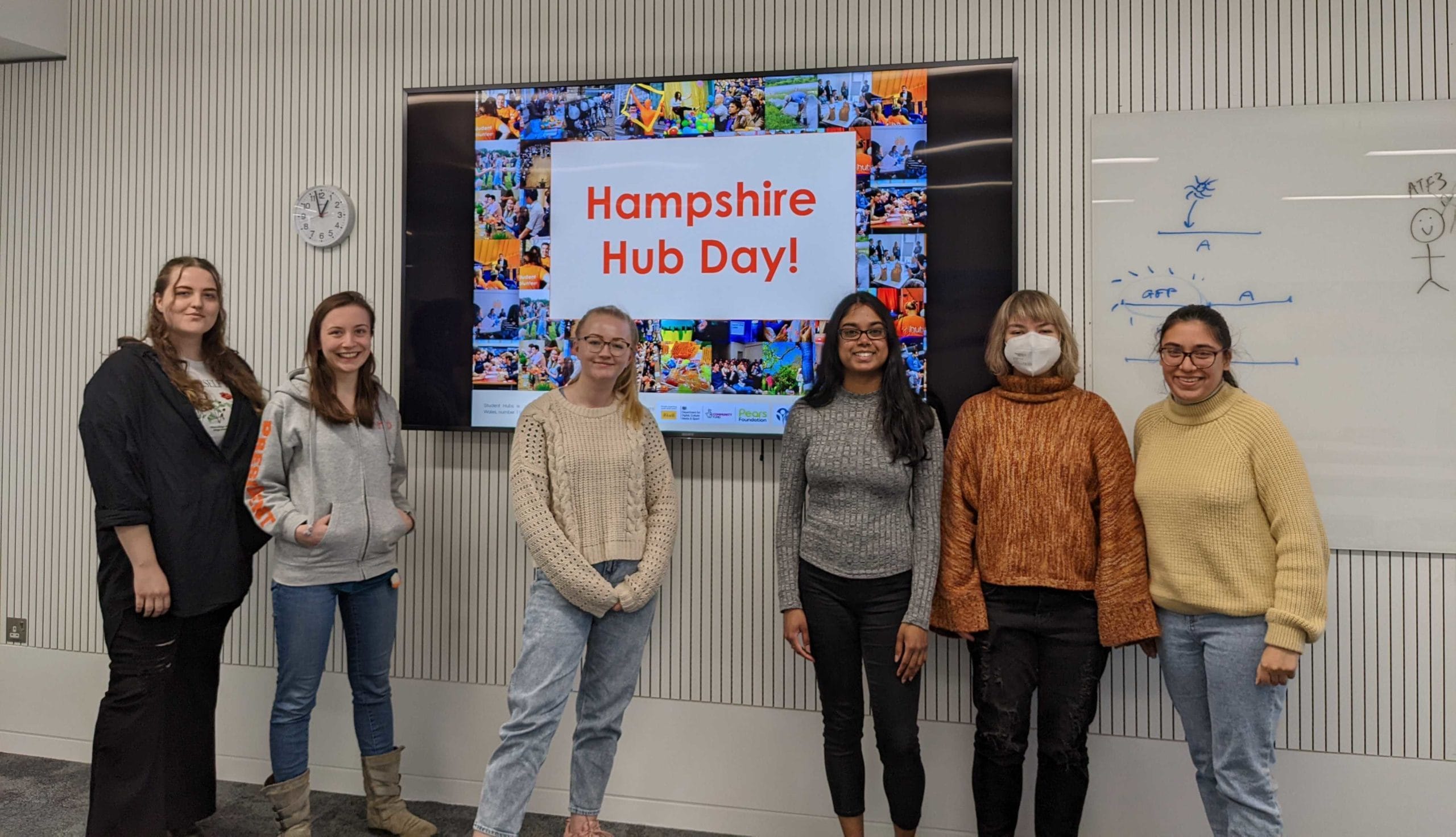 6 individuals stand smiling at the camera in front of a Digital screen showing a slide reading “Hampshire Hub Day”