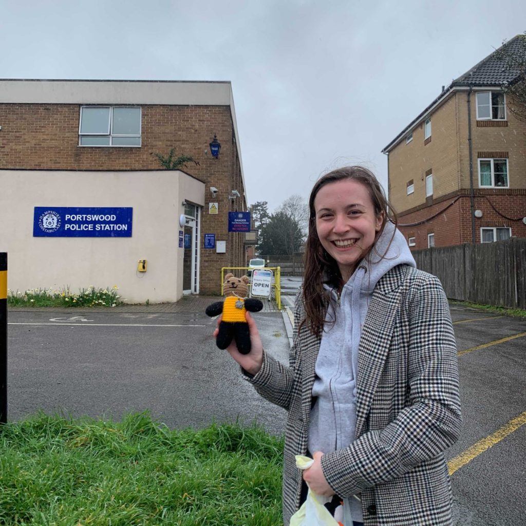 Winchester Hub Programmes Manager, Lydia, stands in front of Portswood Police Station holding a small knitted bear. 