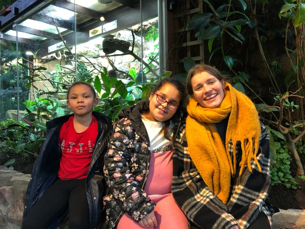 One adult and two children sit smiling at the camera. Behind them are plants.