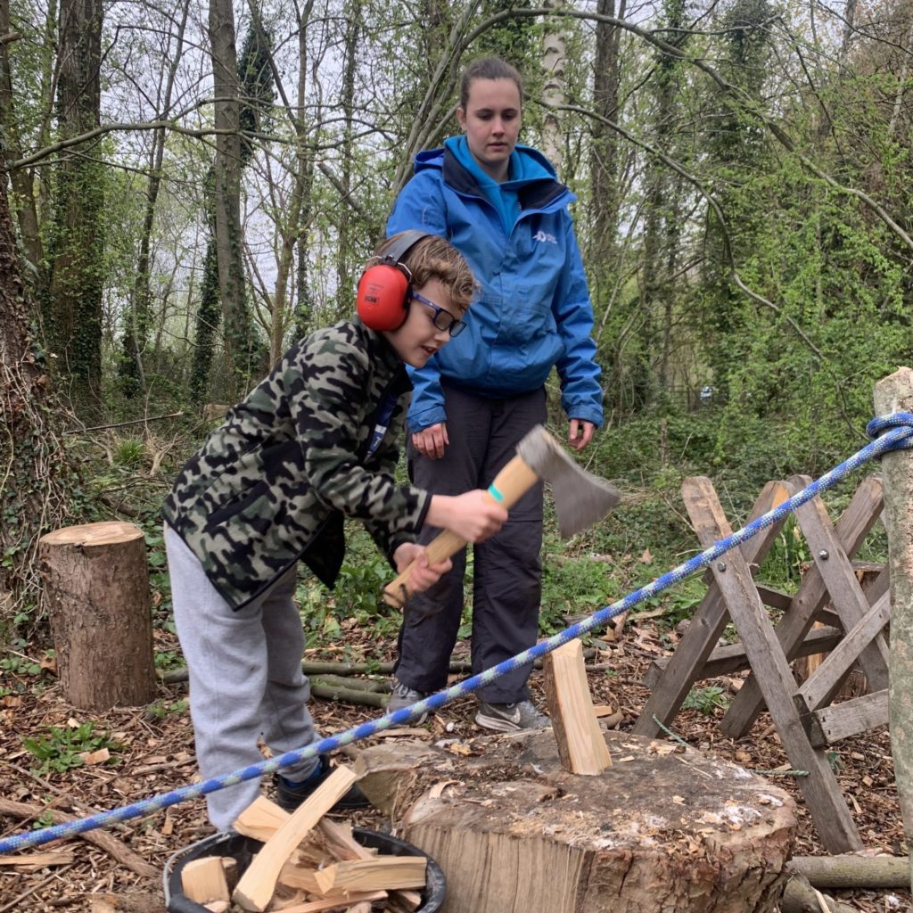 A young person is outside chopping wood supervised by an adult