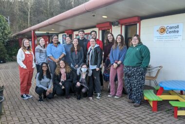 14 members of the Student Hubs team stand outside a community centre smiling at the camera