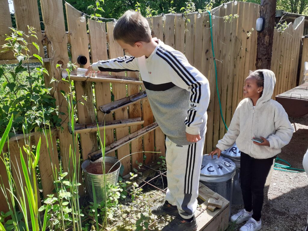 A young person is pouring water into a bamboo shoot that filters into the ground below. Another young person is behind them smiling.