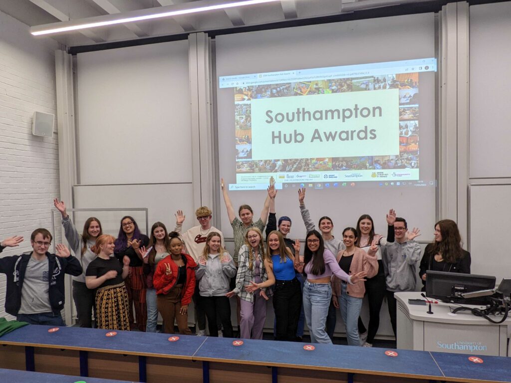 A group of staff and students are stood in front of a projector smiling and waving. On the projector is text "Southampton Hub Awards".