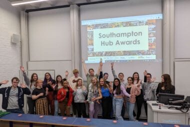 A group of staff and students are stood in front of a projector smiling and waving. On the projector is text "Southampton Hub Awards".