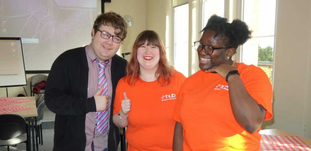 Three individuals are stood smiling at the camera indoors. Two of them are wearing bright orange Hub t-shirts.