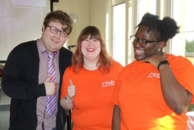Three individuals are stood smiling at the camera indoors. Two of them are wearing bright orange Hub t-shirts.