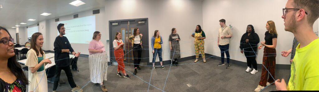 14 members of the Student Hubs staff team stand in a circle with a yarn web between them