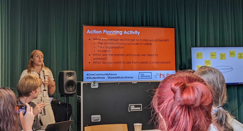 Four individuals look at a presenter. The presenter is stood next to a screen reading 'Action Planning Activity' in front of a dark green curtain.