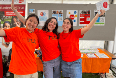 Three volunteers wearing orange t-shirts with the Bristol Hub logo on it, smiling at the camera in front of their Freshers Fayre Stall.