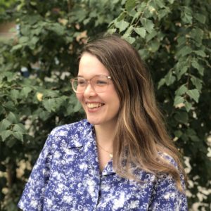 Kingston Hub Programmes Manager, Louise, stands in front of foliage smiling. She is looking slightly to the left.