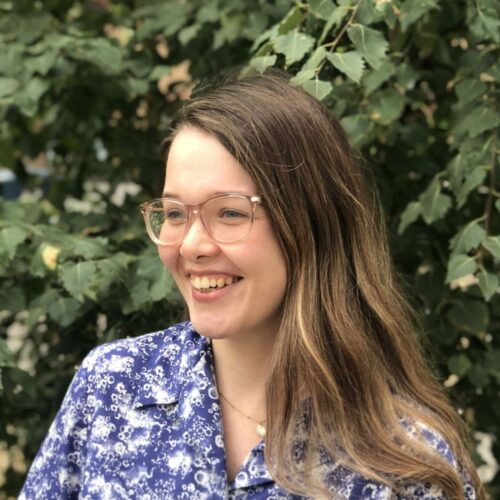 Kingston Hub Programmes Manager, Louise, stands in front of foliage smiling. She is looking slightly to the left.