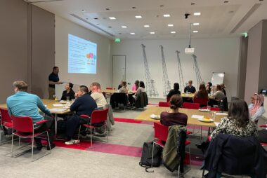 Four tables of individuals sit in a room listening to a presenter