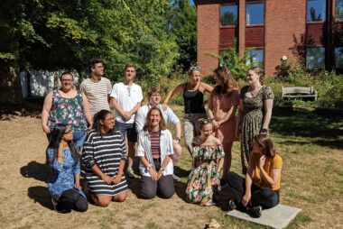 12 members of the Student Hubs team are outside. They are in two rows chatting and smiling at the camera, one stood, and one sat on the grass