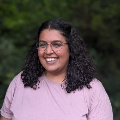 Student Hubs CEO, Simran, stands in front of foliage smiling. She is looking slightly to the left.