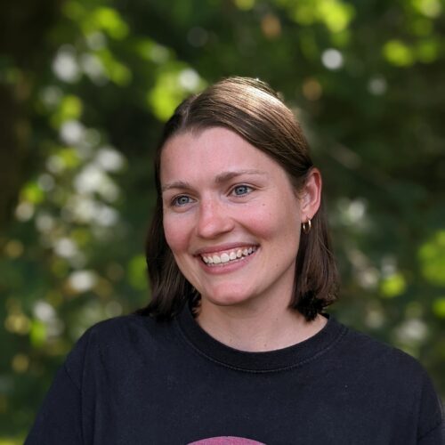 Student Hubs Network Director, Sophie, stands in front of foliage smiling. She is looking slightly to the left.