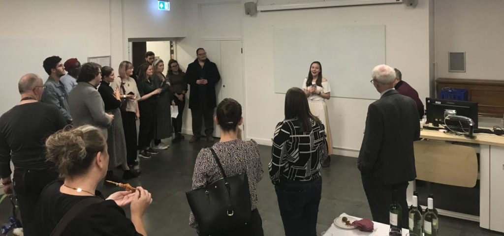 Individuals are stood in a semi circle, listening to a person speaking at the front of a classroom