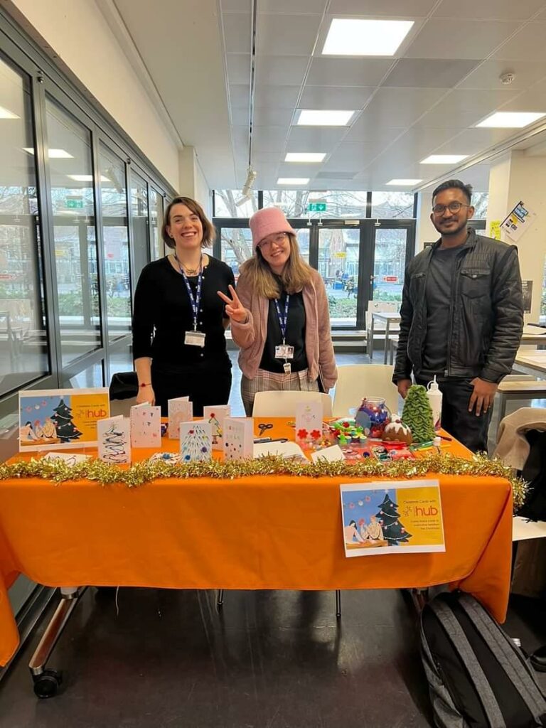 Two staff member and a committee member standing behind a table with orange cloth on with handmade christmas cards on the table.