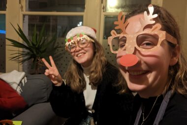 Two staff members looking and smiling at the camera while wearing Christmas glasses one as a Christmas pudding and other as a reindeer.