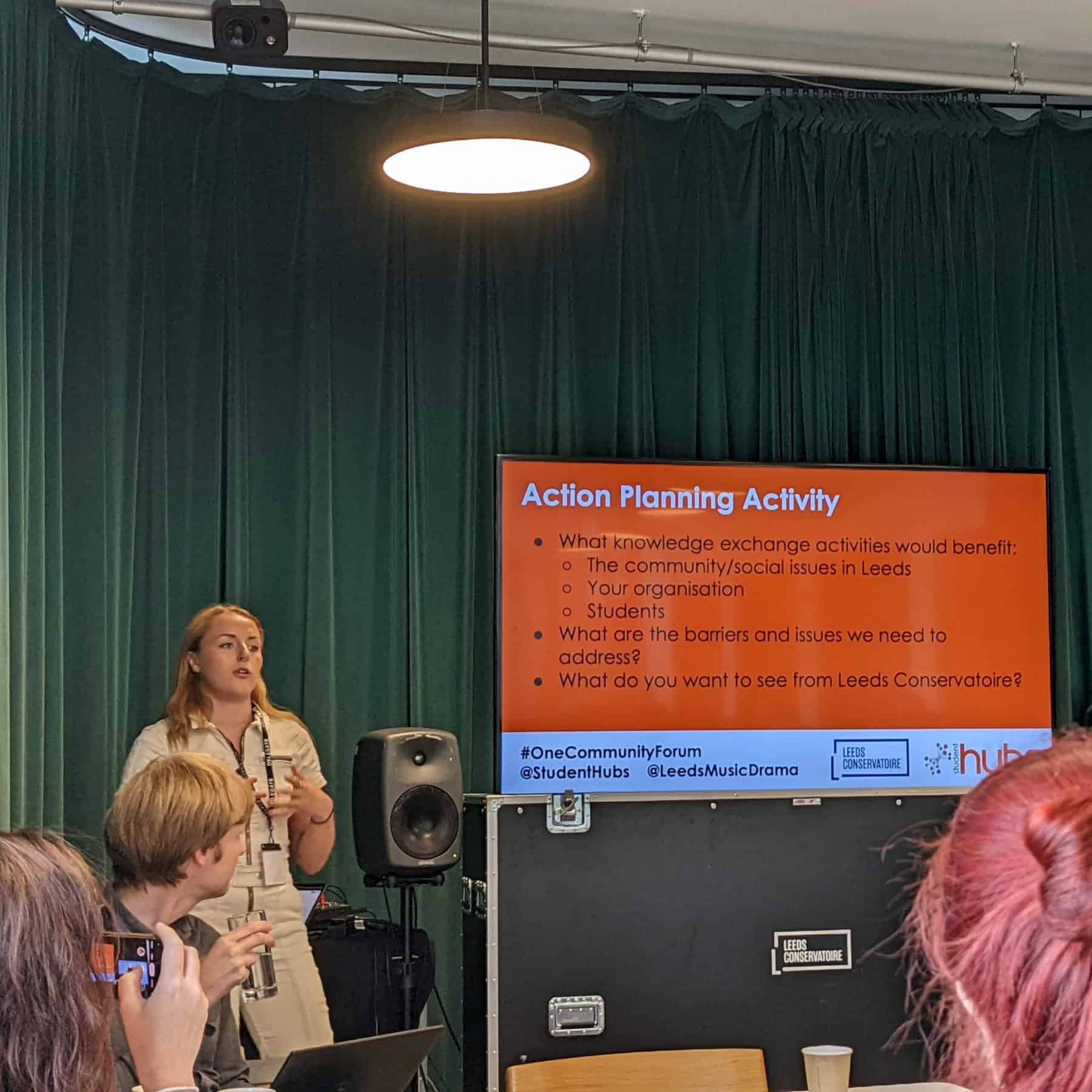 Four individuals look at a presenter. The presenter is stood next to a screen reading 'Action Planning Activity' in front of a dark green curtain.