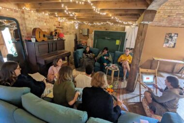 A group of people having a discussion. Half of the group is sat on a blue sofa facing away from the camera, one person is sat on the floor, and everyone else is sat on another sofa. The ceiling is low and there are fairy lights hanging from its wooden beams.